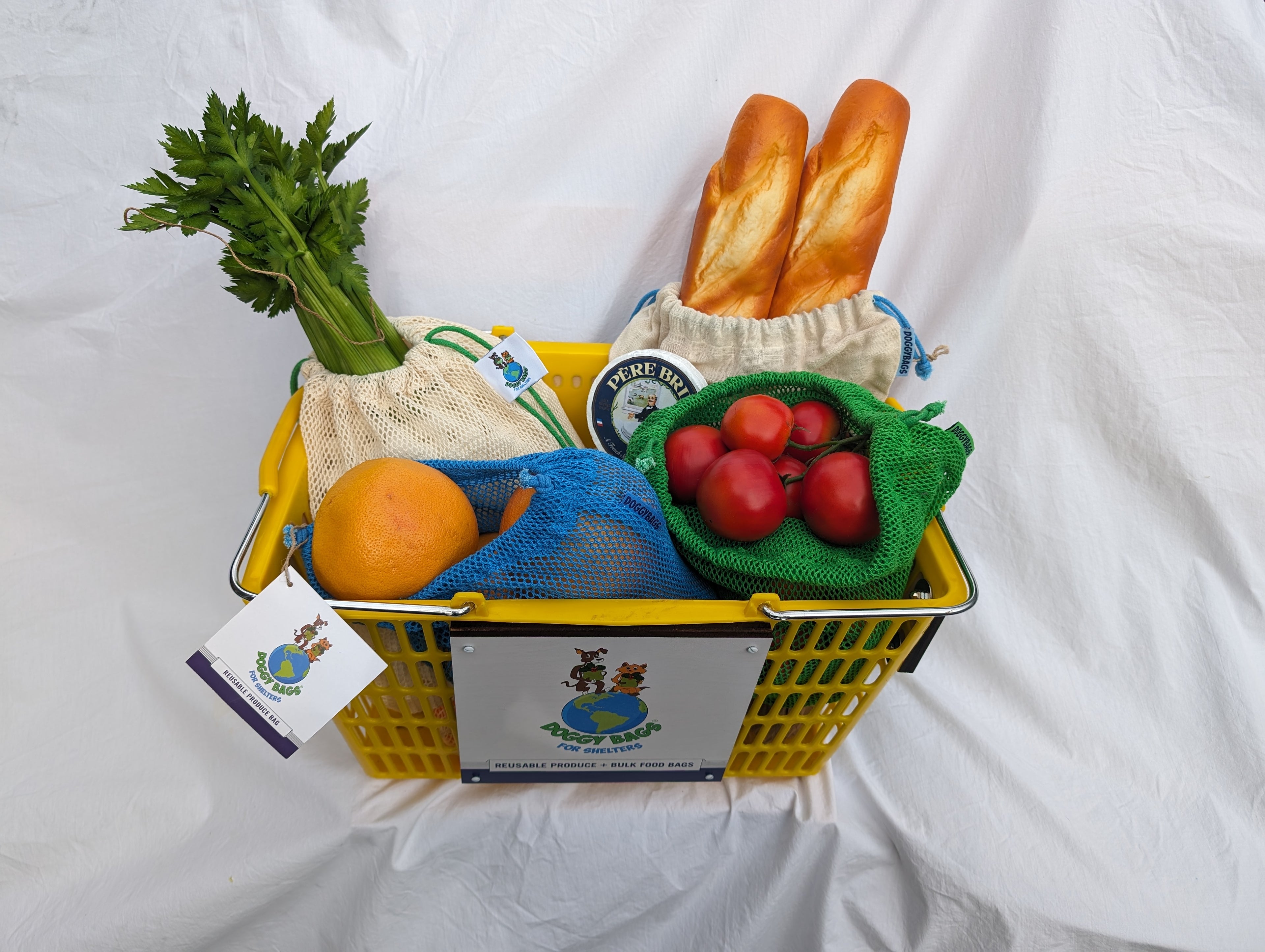 Basket showing bread (2 baguettes) in a bulk food bag, 2 bunches of celery in a natural mesh produce bag, lots of tomatoes in a green produce bag and grapefruit in a blue reusable bag.